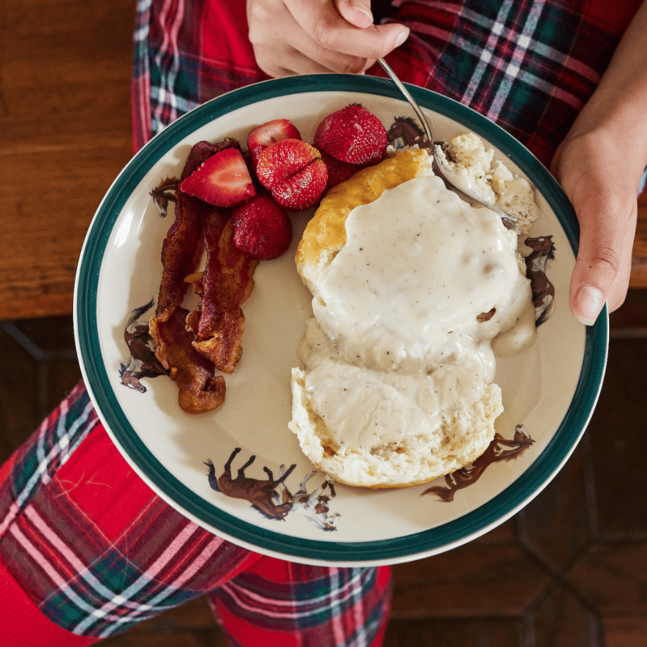 Plate with bacon, strawberries, biscuits and gravy, resting on lap with red plaid pants, hand holding fork