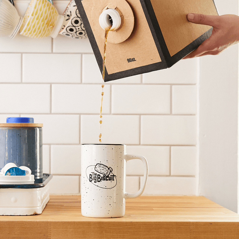 Pouring coffee into white mug with The Big Biscuit logo, white tile in background